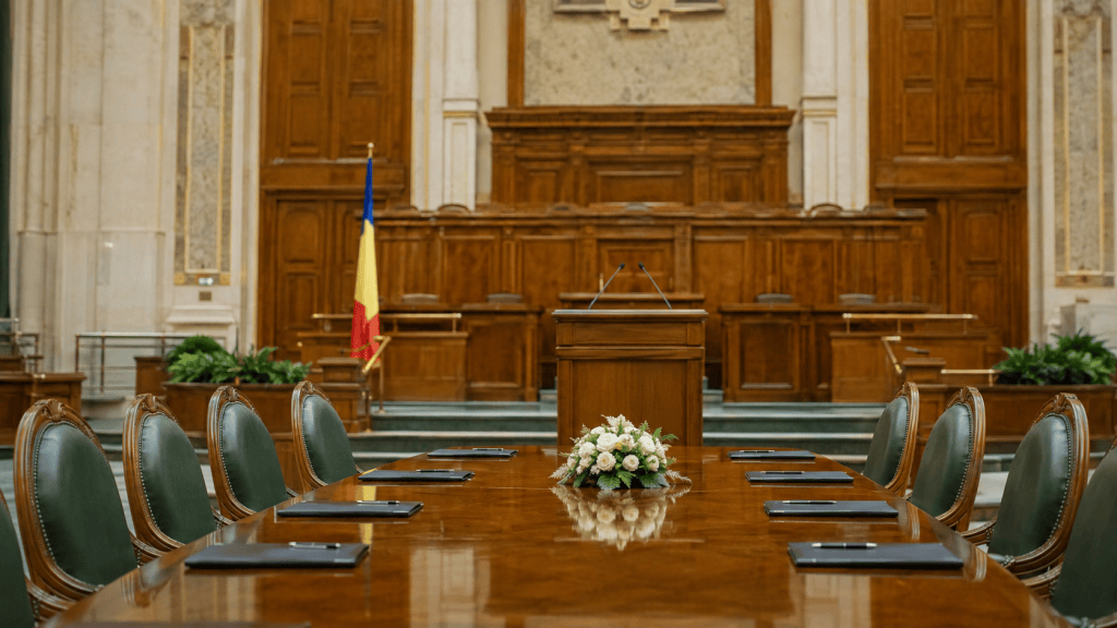 Masă de negocieri și tribună într-o sală instituțională din România, cu drapelul național